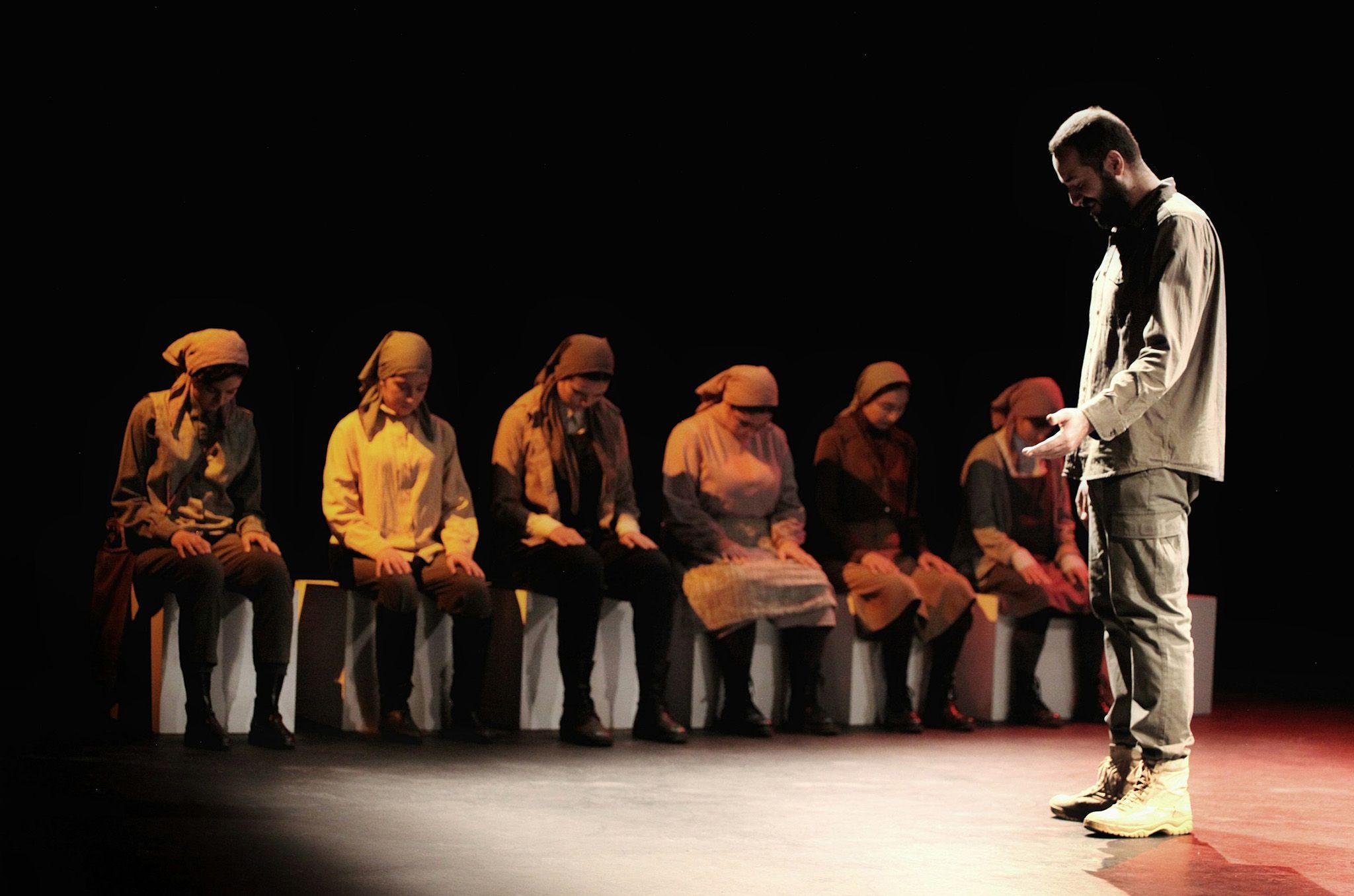 Seven actors on stage, 6 sitting with their heads lowerer and one standing in the spotlight, head lowered.